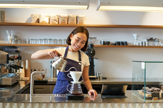Bezpłatne zdjęcie uśmiechnięta azjatycka dziewczyna barista nalewa gorącą wodę z czajnika do parzenia kawy z filtrem stojącym za hrabią