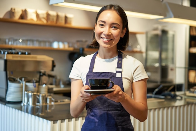 Uśmiechnięta azjatycka dziewczyna barista robi kawę trzymając filiżankę w kawiarnianym fartuchu stojącym w pobliżu hrabiego