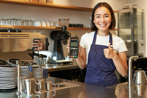Uśmiechnięta azjatycka dziewczyna barista, właścicielka kawiarni w fartuchu, pokazująca czytnik kart płatniczych biorący kontakty