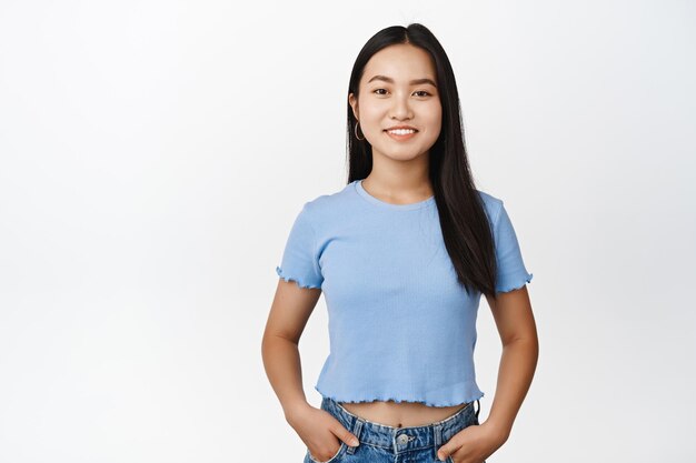 Uśmiechnięta azjatycka dziewczyna stojąca w niebieskiej koszulce, która wygląda pewnie i szczęśliwie w aparacie stojącym na białym tle