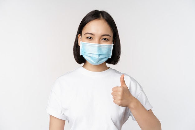 Uśmiechnięta Azjatycka dziewczyna w masce medycznej pokazująca kciuki w górę, aprobuje coś dobrego pochwały i komplementuje firmę stojącą na białym tle
