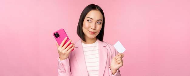 Uśmiechnięta azjatycka kobieta korporacyjna w garniturze myśląca, trzymająca smartfona i kartę kredytową, planuje kupić coś online na różowym tle telefonu komórkowego