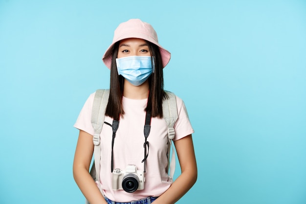 Uśmiechnięta azjatycka kobieta z medyczną maską na twarz podróżująca podczas pandemii stojąc z aparatem fotograficznym...