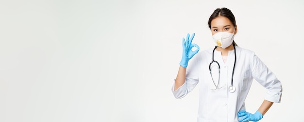 Bezpłatne zdjęcie uśmiechnięta azjatycka lekarka, lekarka, pokazuje znak, że nosi respirator po złapaniu choroby covid s