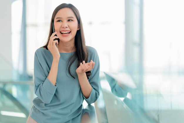 Uśmiechnięta azjatycka nastolatka ręka za pomocą smartfona rozmowa z bliskim przyjacielem w centrum handlowym lub na korytarzu uczelni wesoły śmiech uśmiech pozytywne nastawienie emocjamłoda azja kobieta smartfon