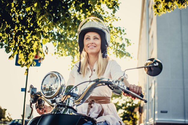 Uśmiechnięta blond dziewczyna w kasku moto siedzi na klasycznym skuterze moto pod zielonym drzewem.