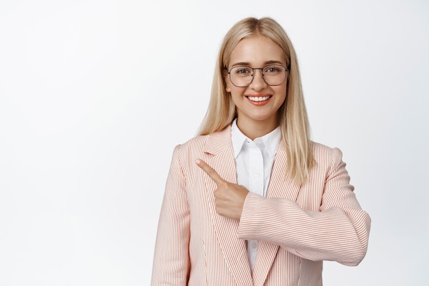 Uśmiechnięta blond kobieta w okularach wskazujący palec w lewo, w okularach i różowym kolorze na białym.