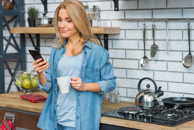 Uśmiechnięta blondynki młoda kobieta trzyma filiżankę kawy patrzeje telefon komórkowego