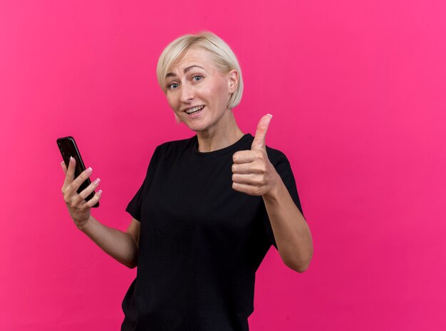 Uśmiechnięta blondynki słowiańska kobieta w średnim wieku, trzymając telefon komórkowy pokazując kciuk do góry na białym tle na szkarłatnej ścianie z miejsca na kopię