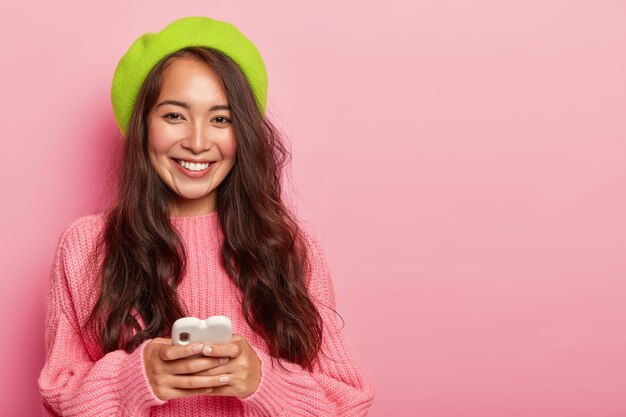 Uśmiechnięta brunetka kobieta z długimi włosami, nosi jasnozielony beret i duży sweter, trzyma nowoczesny telefon komórkowy podłączony do bezprzewodowego internetu