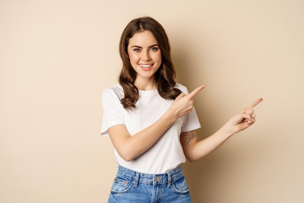 Bezpłatne zdjęcie uśmiechnięta brunetka w koszulce, wskazująca palcami w prawo, pokazująca ofertę promocyjną lub reklamę, pokazująca baner, stojąca na beżowym tle