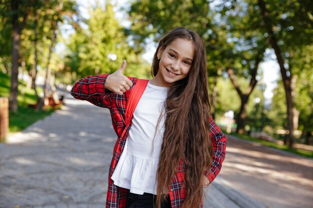 Uśmiechnięta brunetki młoda dziewczyna pozuje w parku i pokazuje kciuk up