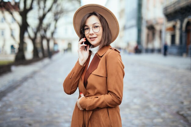 Uśmiechnięta dama w szerokim kapeluszowym mówieniu na smartphone jesieni ulicie