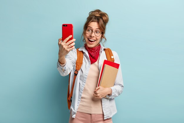 Uśmiechnięta, dobrze wyglądająca kobieta robi selfie przez telefon komórkowy, będąc w dobrym nastroju, lubi wolny czas po zajęciach, nosi czerwoną chustkę na szyi