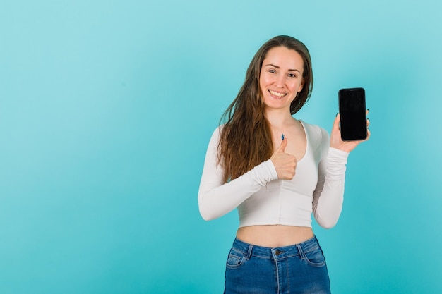 Uśmiechnięta dziewczyna pokazuje pomysł na makieta z telefonem komórkowym i pokazuje doskonały gest drugą ręką na niebieskim tle