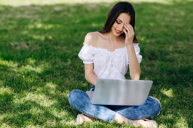 Uśmiechnięta dziewczyna siedzi w parku, wpisując na swoim laptopie