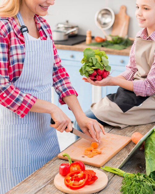 Uśmiechnięta dziewczyna trzyma rzodkiewkę w ręku pomaga jej matka cięcia marchew z nożem na desce do krojenia