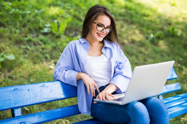 Uśmiechnięta dziewczyna w niebieskiej koszulce siedzieć na ławce w parku i używać swojego nowego laptopa