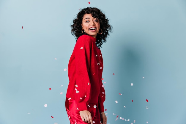 Uśmiechnięta ekstatyczna dziewczyna pozuje z konfetti Studio strzał młodej kobiety rasy mieszanej w swobodnym czerwonym swetrze