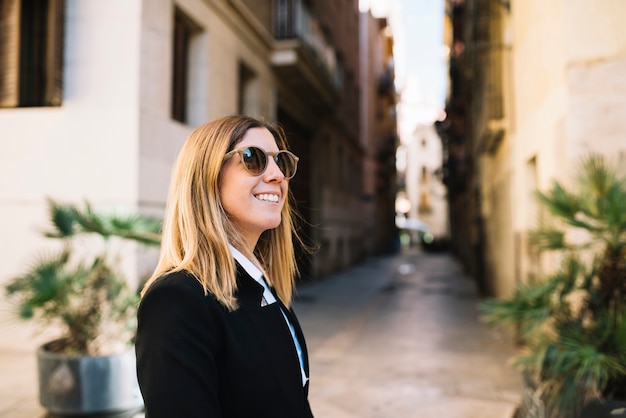 Uśmiechnięta elegancka młoda kobieta z okularami przeciwsłonecznymi na wąskiej ulicie