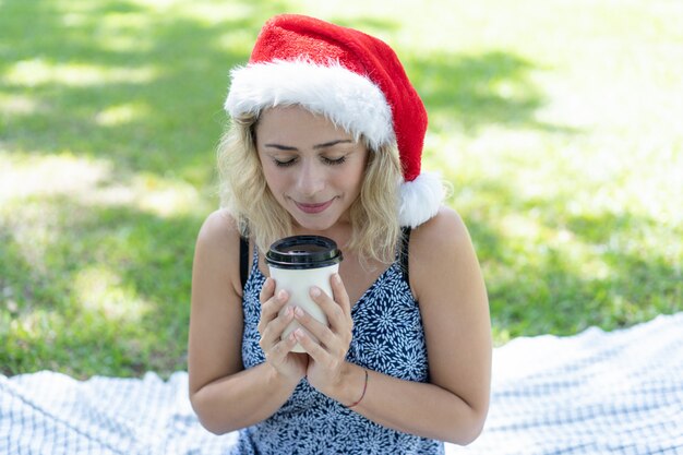 Uśmiechnięta kobieta jest ubranym Santa kapelusz i cieszy się kawę w parku