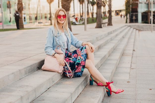 uśmiechnięta kobieta siedzi na schodach na ulicy miasta w stylowej drukowanej spódnicy i dżinsowej kurtce oversize ze skórzanym plecakiem na sobie różowe okulary przeciwsłoneczne