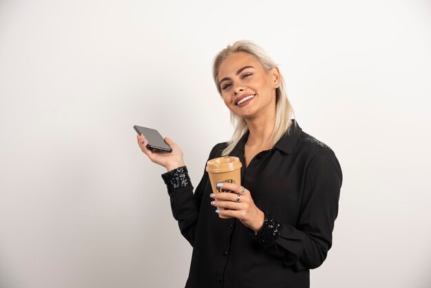 Uśmiechnięta kobieta stojąca z telefonem komórkowym i filiżanką kawy. Wysokiej jakości zdjęcie