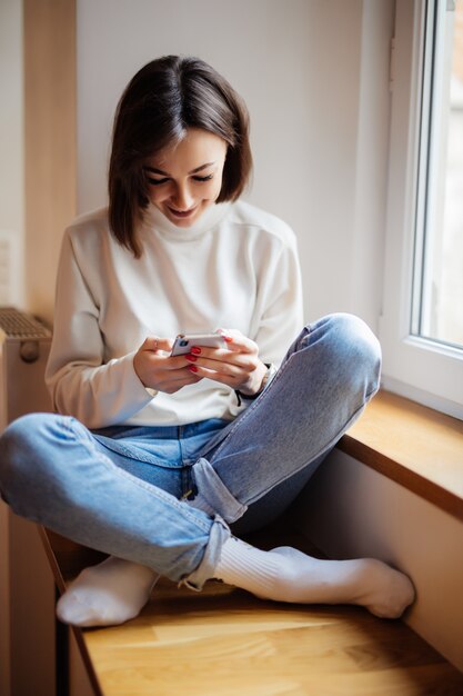 Uśmiechnięta kobieta w białym pulowerze siedzi na parapecie i pisze wiadomości na smartphone w domu