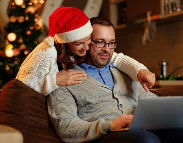 Uśmiechnięta kobieta w czapce Mikołaja i pulchny pozytywny mężczyzna korzystający z laptopa na kanapie w pokoju ze świąteczną dekoracją.