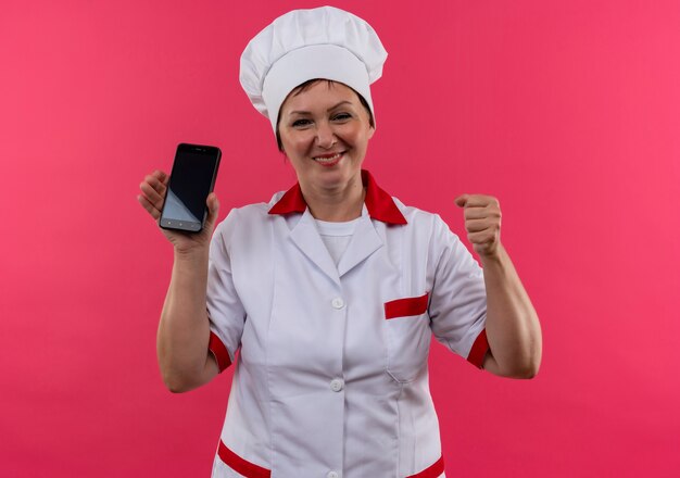 Uśmiechnięta kobieta w średnim wieku kucharz w mundurze szefa kuchni trzymając telefon pokazujący tak gest