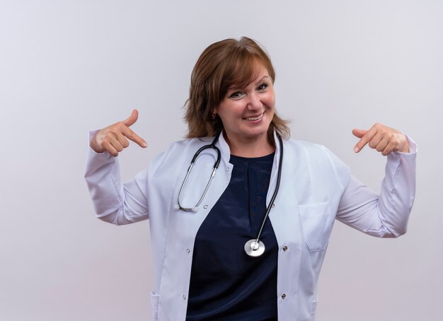 Uśmiechnięta kobieta w średnim wieku lekarz ubrana w medyczny szlafrok i stetoskop, wskazując palcami na siebie na odizolowanej białej ścianie