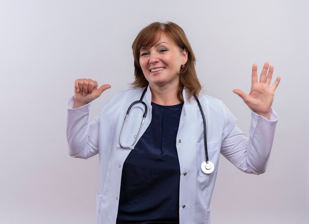 Uśmiechnięta kobieta w średnim wieku lekarz ubrana w szlafrok i stetoskop, wskazując palcem na siebie i pokazując pięć na odizolowanej białej ścianie