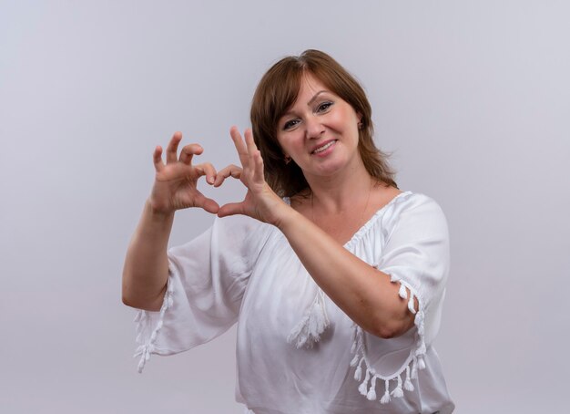 Uśmiechnięta kobieta w średnim wieku pokazująca znak serca z rękami na odosobnionej białej ścianie