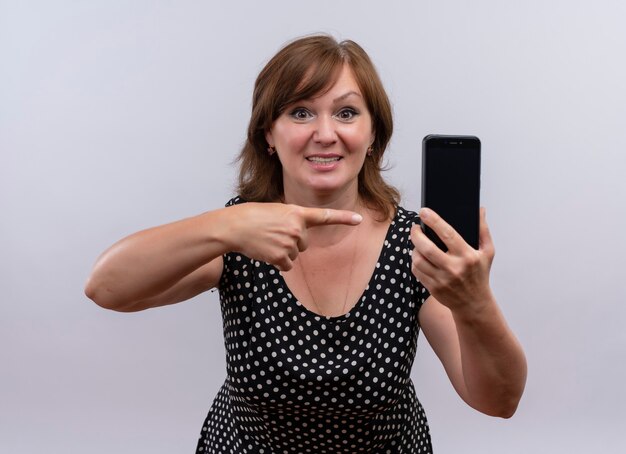 Uśmiechnięta kobieta w średnim wieku trzyma telefon komórkowy i wskazuje palcem na niego na odosobnionej białej ścianie