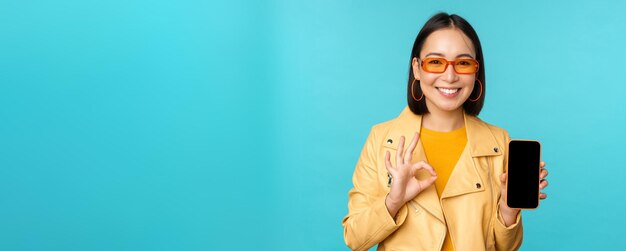Uśmiechnięta koreańska kobieta pokazująca aplikację na telefon komórkowy z interfejsem aplikacji na smartfona, polecającą na telefonie komórkowym stojącym na niebieskim tle