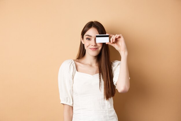 Uśmiechnięta ładna dziewczyna pokazuje plastikową kartę kredytową nad oczami i wygląda na zadowoloną, stojąc na beżowym bac...