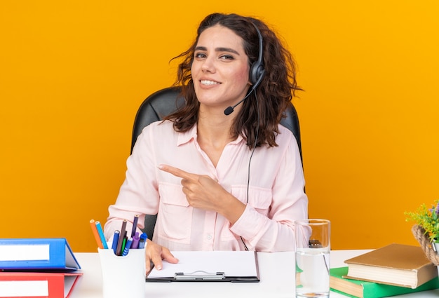 Uśmiechnięta ładna kaukaska operatorka call center na słuchawkach siedząca przy biurku z narzędziami biurowymi wskazującymi na bok na pomarańczowej ścianie