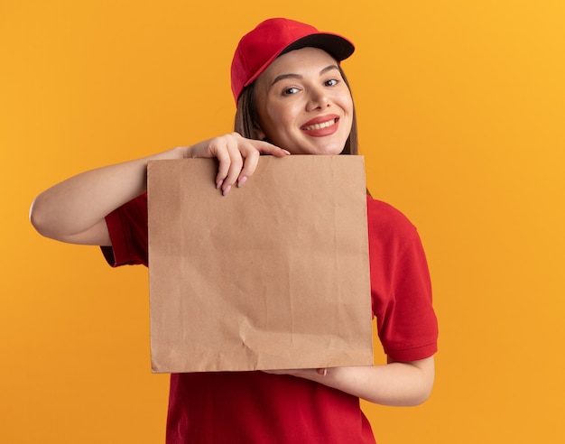 Uśmiechnięta ładna kobieta dostawy w mundurze posiada pakiet papieru na białym tle na pomarańczowej ścianie z miejsca na kopię