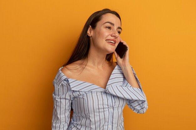 Uśmiechnięta ładna kobieta rozmawia przez telefon, patrząc na bok na białym tle na pomarańczowej ścianie
