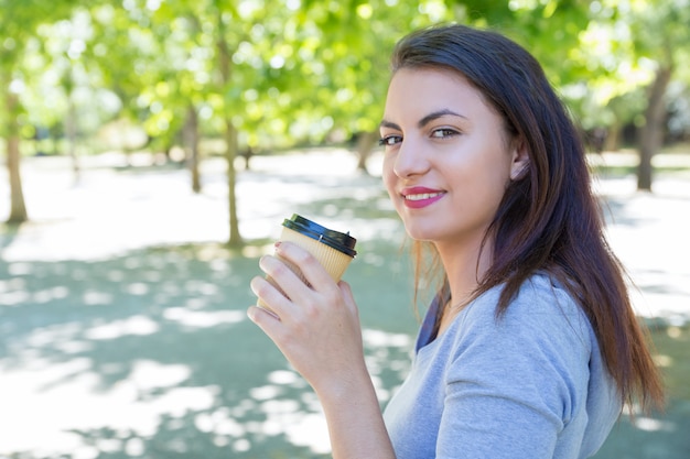 Uśmiechnięta ładna młoda kobieta pije kawę w parku