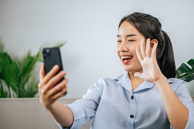 Uśmiechnięta ładna nastolatka siedząca na kanapie i używająca smartfona do wideorozmowy lub selfie podczas kwarantanny samoizolacji covid-19 w domu