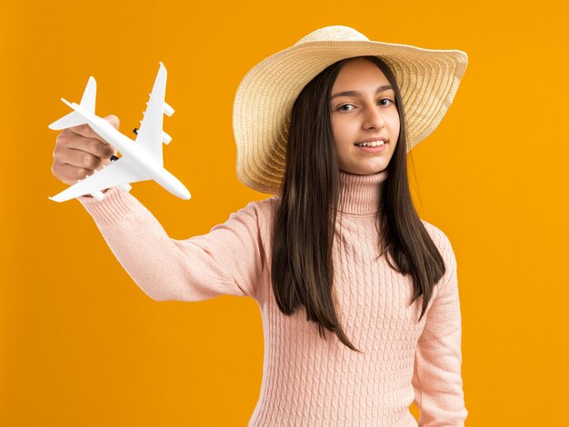 Uśmiechnięta ładna nastolatka w kapeluszu plażowym trzymająca model samolotu na pomarańczowej ścianie