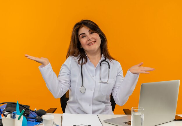 Uśmiechnięta lekarka w średnim wieku ubrana w szlafrok medyczny i stetoskop siedząca przy biurku ze schowkiem na narzędzia medyczne i laptopem pokazująca puste ręce na białym tle