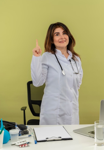 Uśmiechnięta lekarka w średnim wieku w szlafroku medycznym i stetoskopie stojąca za biurkiem z narzędziami medycznymi i laptopem podnoszącym palec, trzymając rękę za plecami odizolowaną