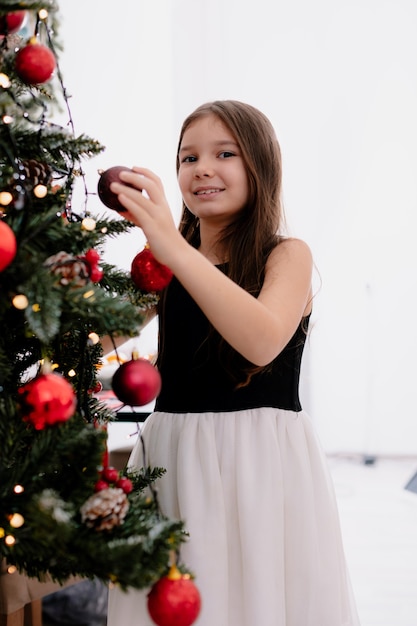 Uśmiechnięta mała dziewczynka w domu w czasie świąt dekoruje choinkę w salonie trzymając bombkę bożonarodzeniową