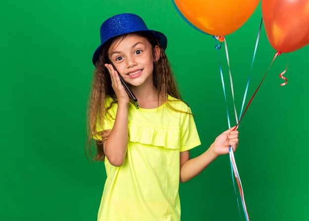 uśmiechnięta mała kaukaska dziewczynka w niebieskim kapeluszu imprezowym trzymająca balony z helem i rozmawiająca przez telefon odizolowany na zielonej ścianie z kopią przestrzeni