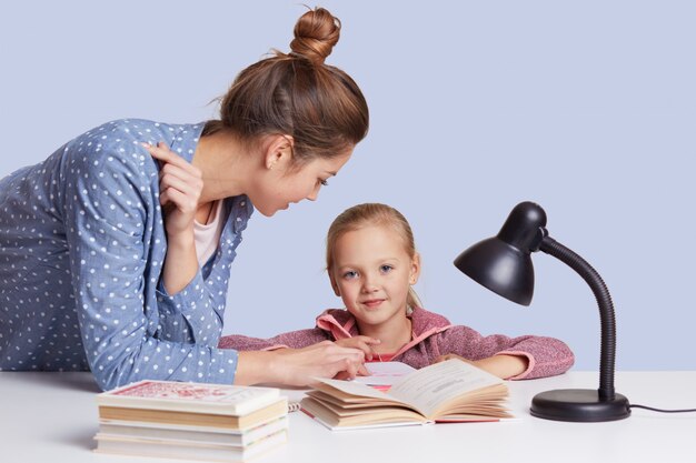 Uśmiechnięta mała powabna dziewczyna siedzi przy stołem, a jej matka pomaga jej odrabiać zadanie domowe, próbuje razem nauczyć się wiersza, używa lampki do czytania dla dobrego widzenia, na białym tle.