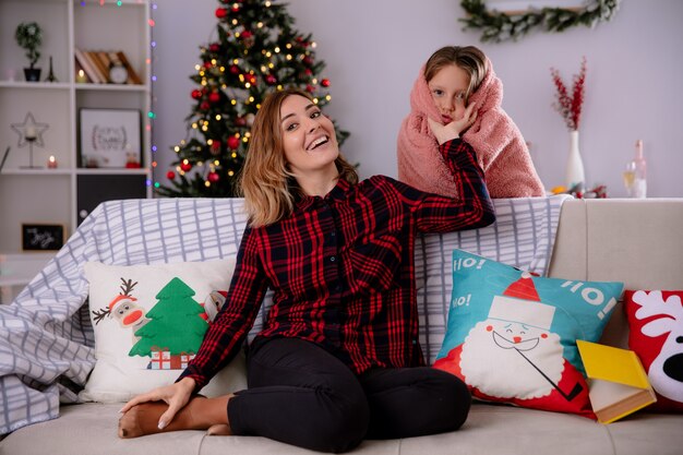 Uśmiechnięta matka siedząca na kanapie trzyma policzki córki zawinięte w koc, ciesząc się świątecznym czasem w domu