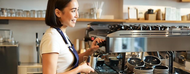 Bezpłatne zdjęcie uśmiechnięta młoda azjatycka barista używająca parownika do kawy, parzącego mleko do zamówienia cappuccino