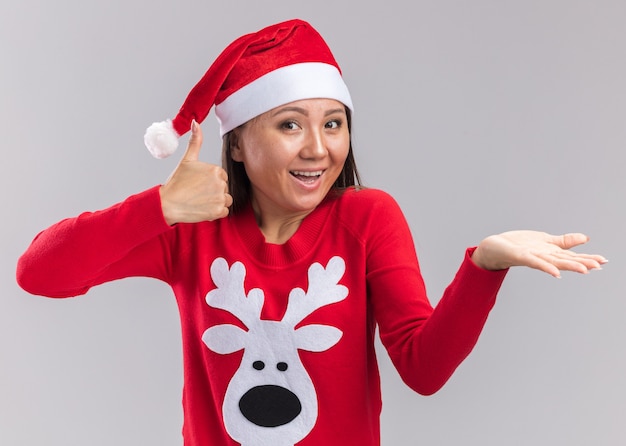 Uśmiechnięta młoda azjatycka dziewczyna ubrana w świąteczny kapelusz ze swetrem pokazując kciuk do góry na białym tle